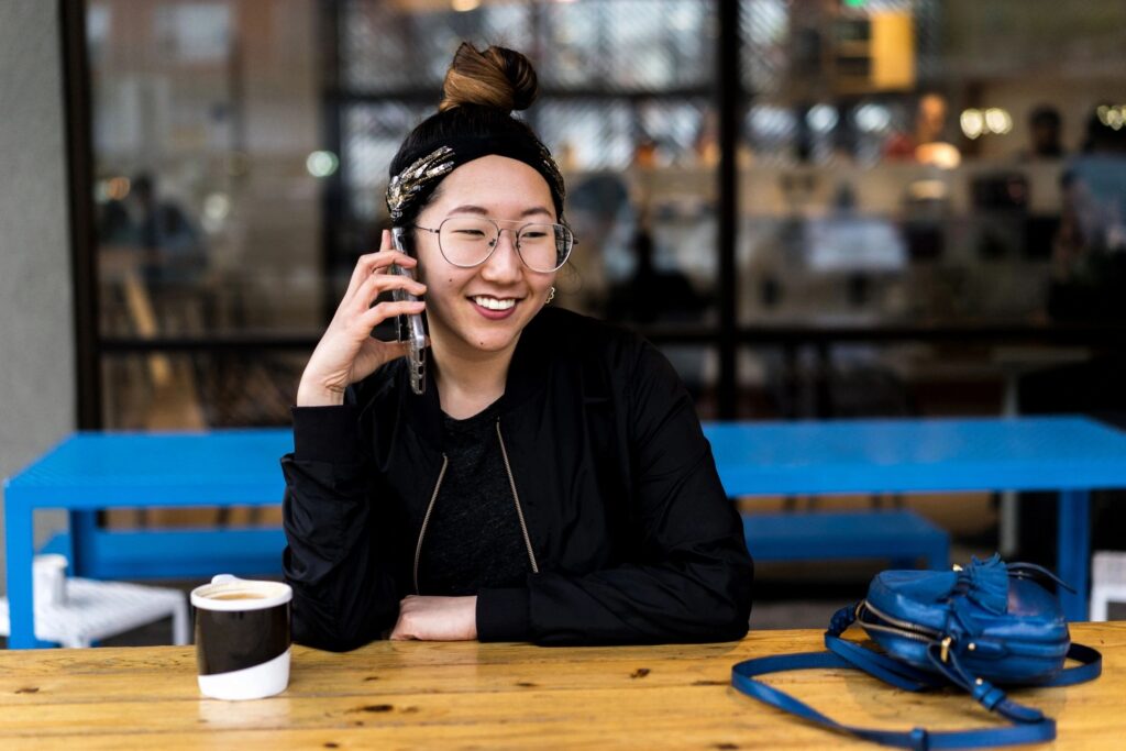 Smiling person with a cell phone to her ear, a coffee, and a restaurant behind her.